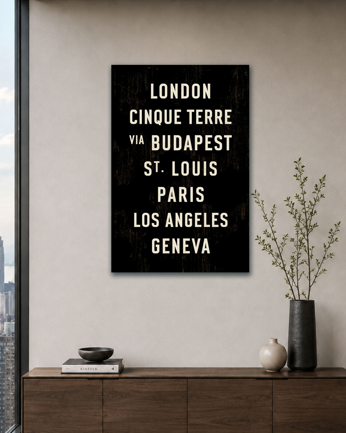 Custom canvas wall art print with white text listing major cities (London, Cinco Terre, via Budapest, St. Louis, Paris, Los Angeles, Geneva) displayed on a beige wall above a wooden console table with decorative vases.