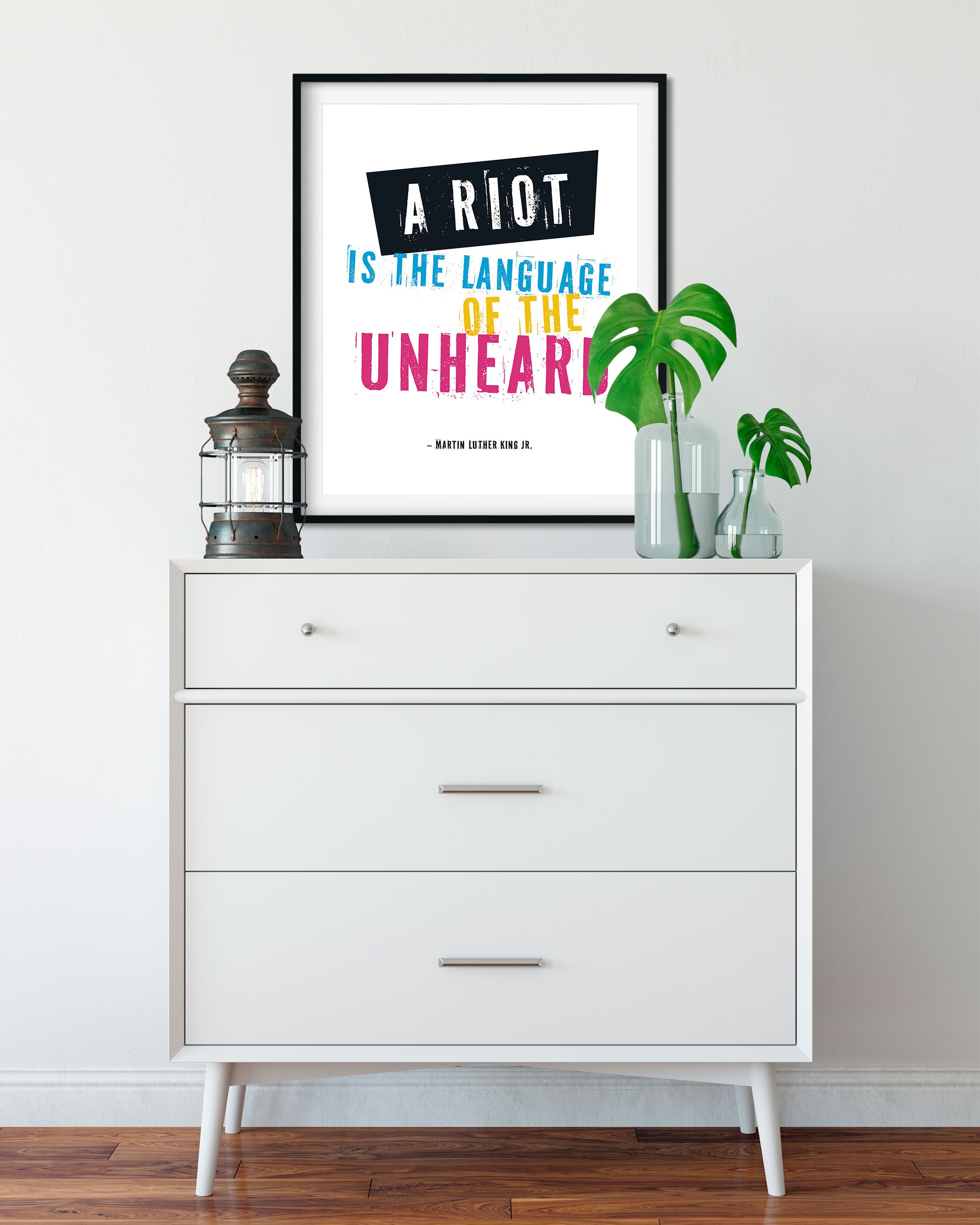 A framed social justice print with the quote 'A RIOT IS THE LANGUAGE OF THE UNHEARD' - a quote by Martin Luther King Jr., is displayed above a white dresser in a styled interior setting with a lantern and plant props.