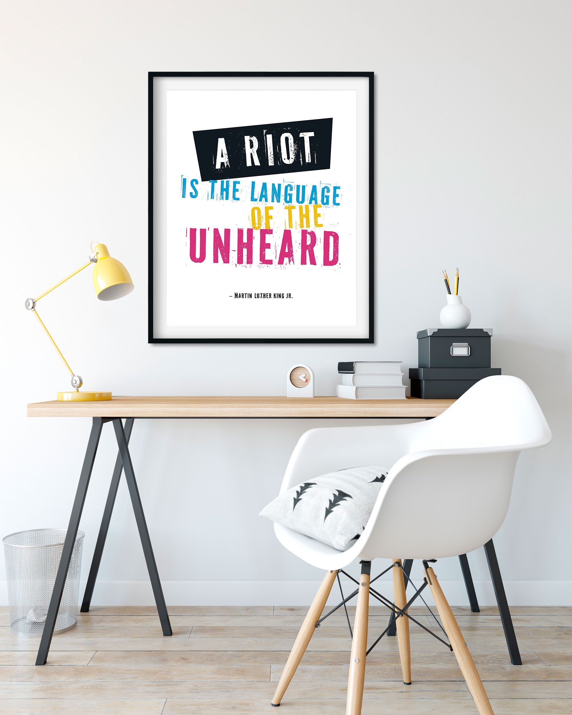 A framed protest poster wall art print hangs above a desk in a bright home office. The print reads “A RIOT IS THE LANGUAGE OF THE UNHEARD” with black, white, blue, yellow, and pink lettering.