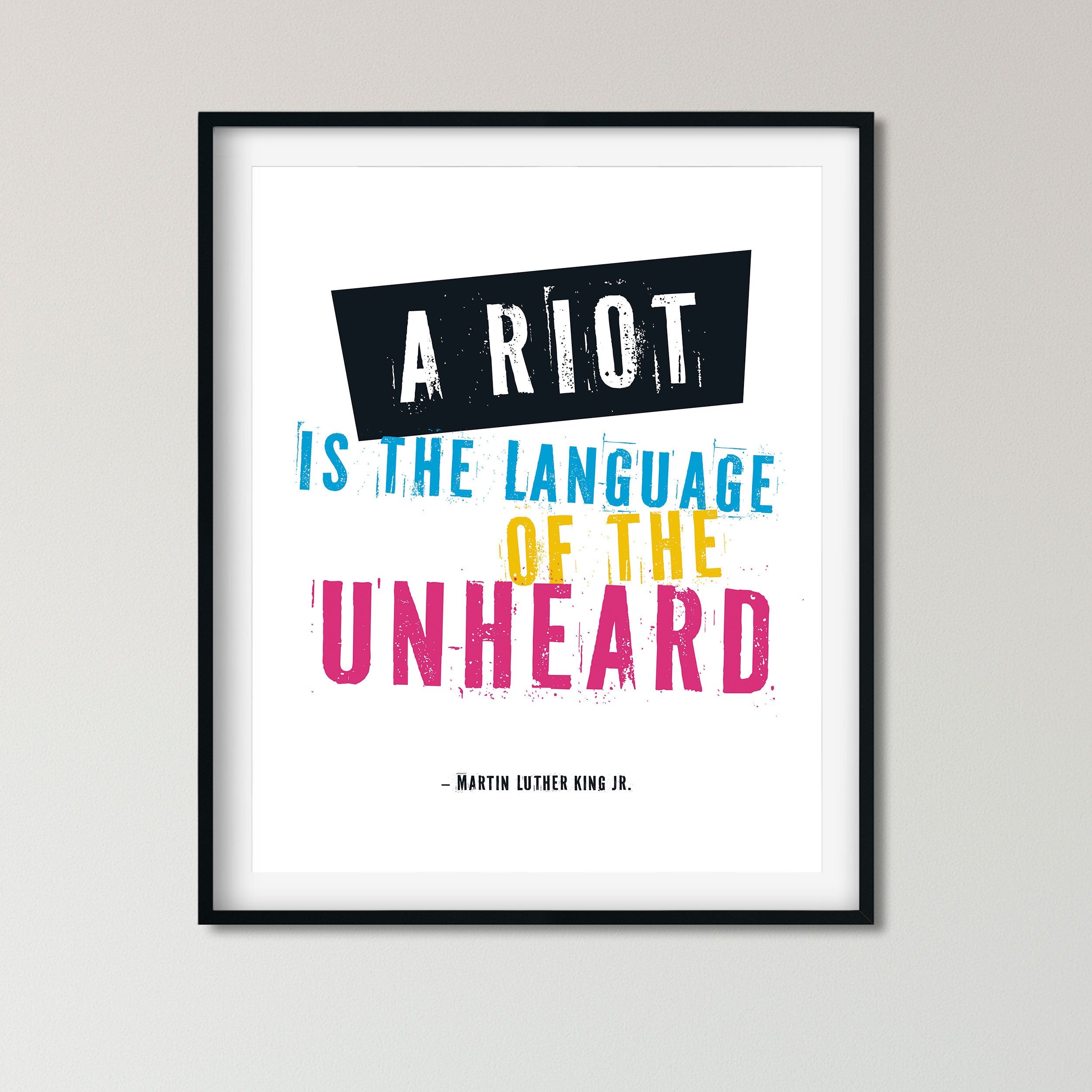 A framed typographic wall art print featuring the quote 'A RIOT IS THE LANGUAGE OF THE UNHEARD' attributed to Martin Luther King Jr., with bold white, blue, yellow, and pink lettering on a white background and a black frame.