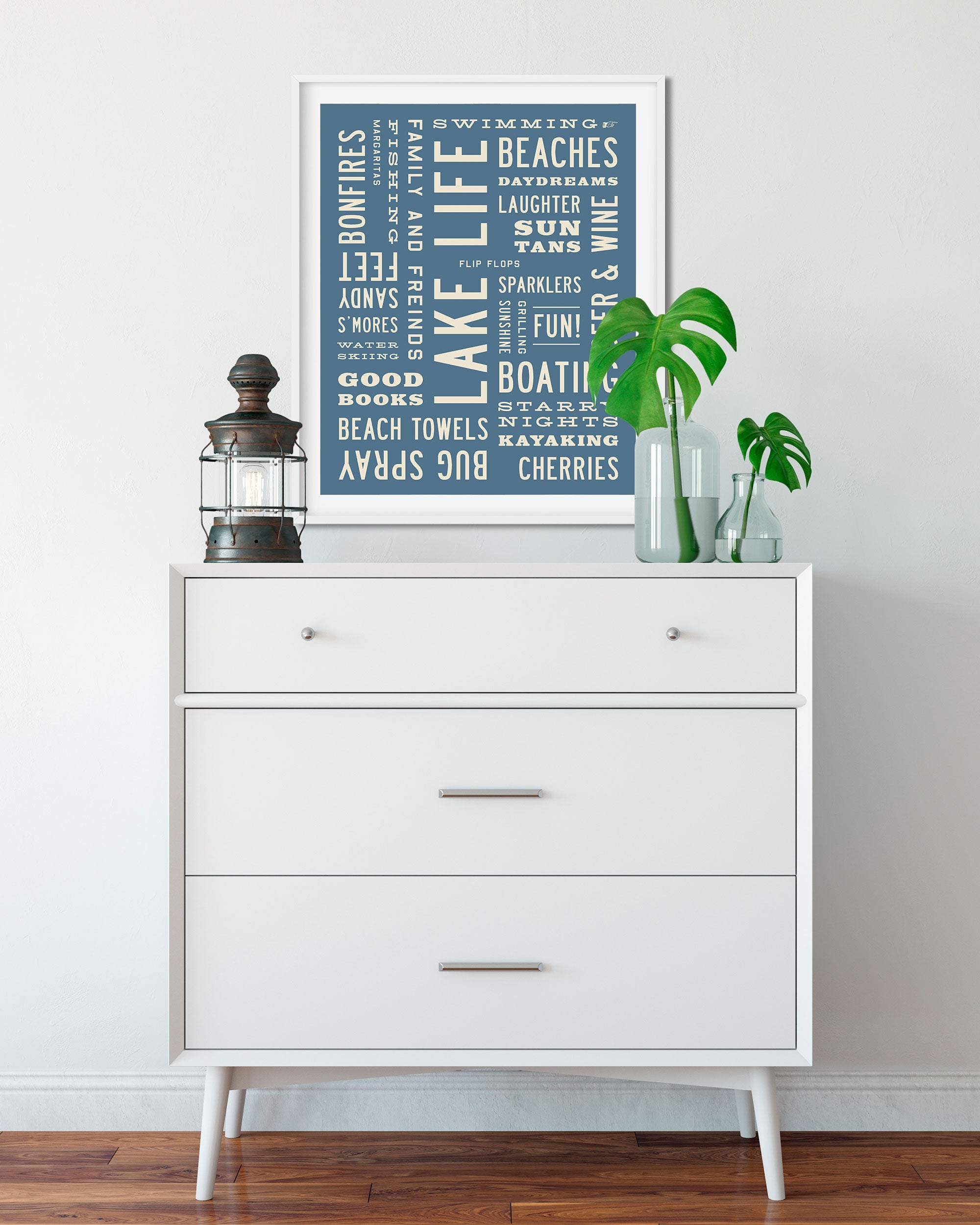 A framed typography art print featuring beach-themed words on a slate blue background, styled on top of a white three-drawer dresser with a lantern and plants in a bright room.