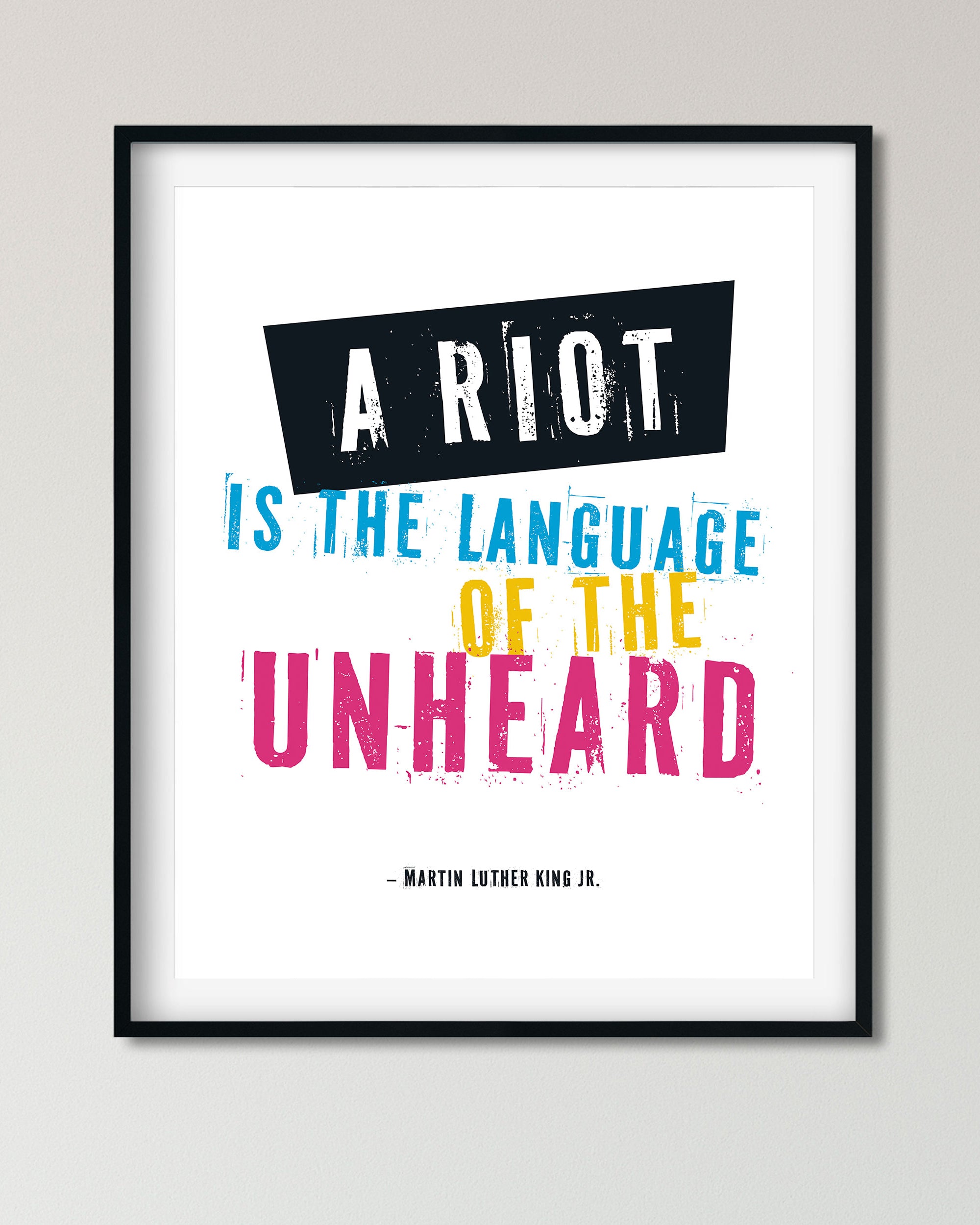 Framed typographic wall art featuring a Martin Luther King Jr. quote: 'A RIOT IS THE LANGUAGE OF THE UNHEARD' in white, blue, yellow, and pink lettering on a white background, shown in a black frame against a light gray wall.