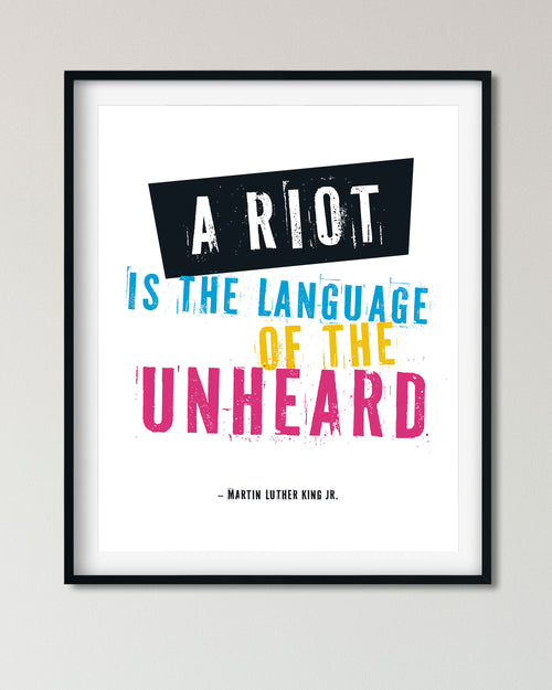 Framed typographic wall art featuring a Martin Luther King Jr. quote: 'A RIOT IS THE LANGUAGE OF THE UNHEARD' in white, blue, yellow, and pink lettering on a white background, shown in a black frame against a light gray wall.